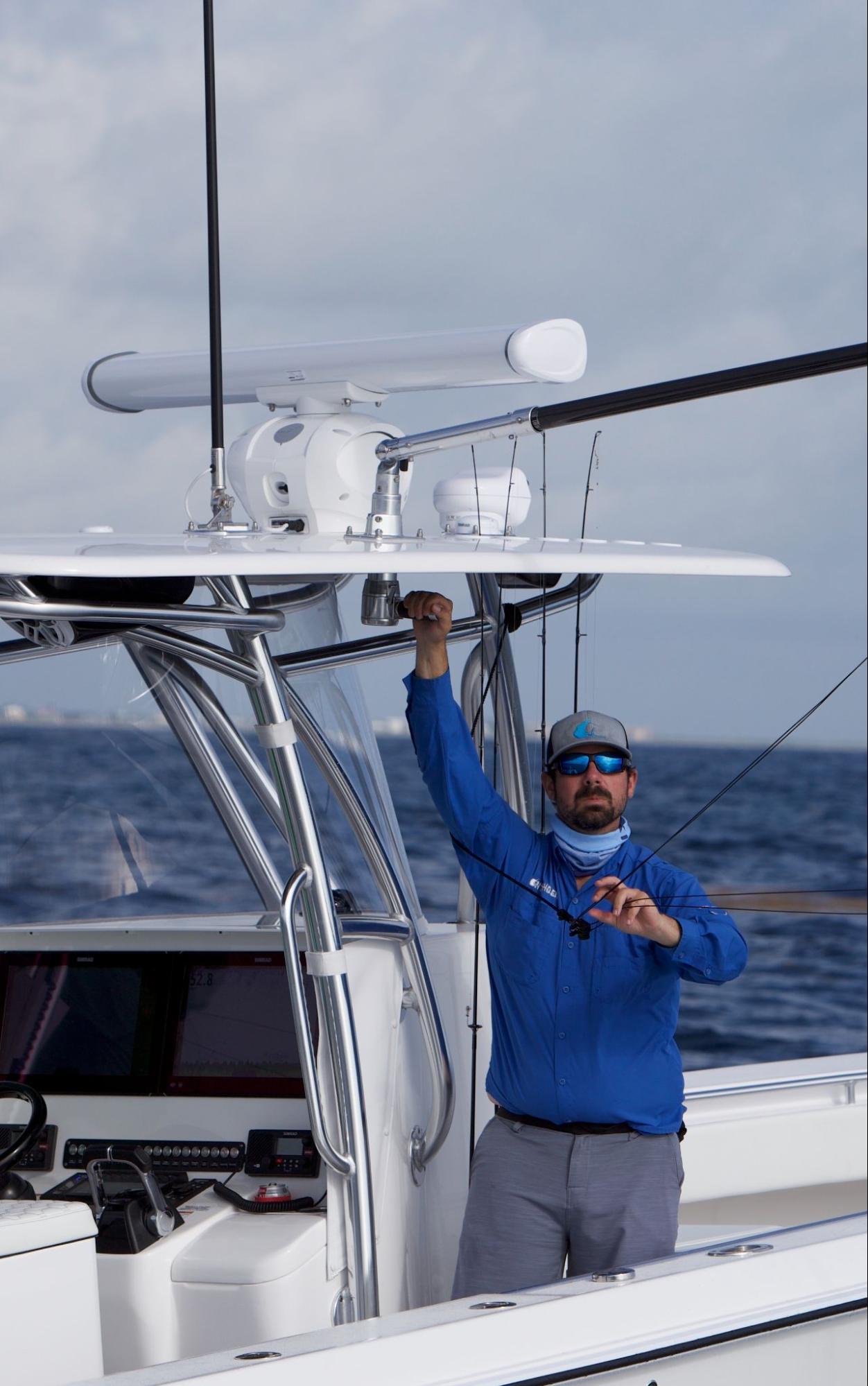 A man in a blue shirt and sunglasses stands on a boat, holding onto the boat's frame with one hand and equipment in the other, with the ocean in the background.