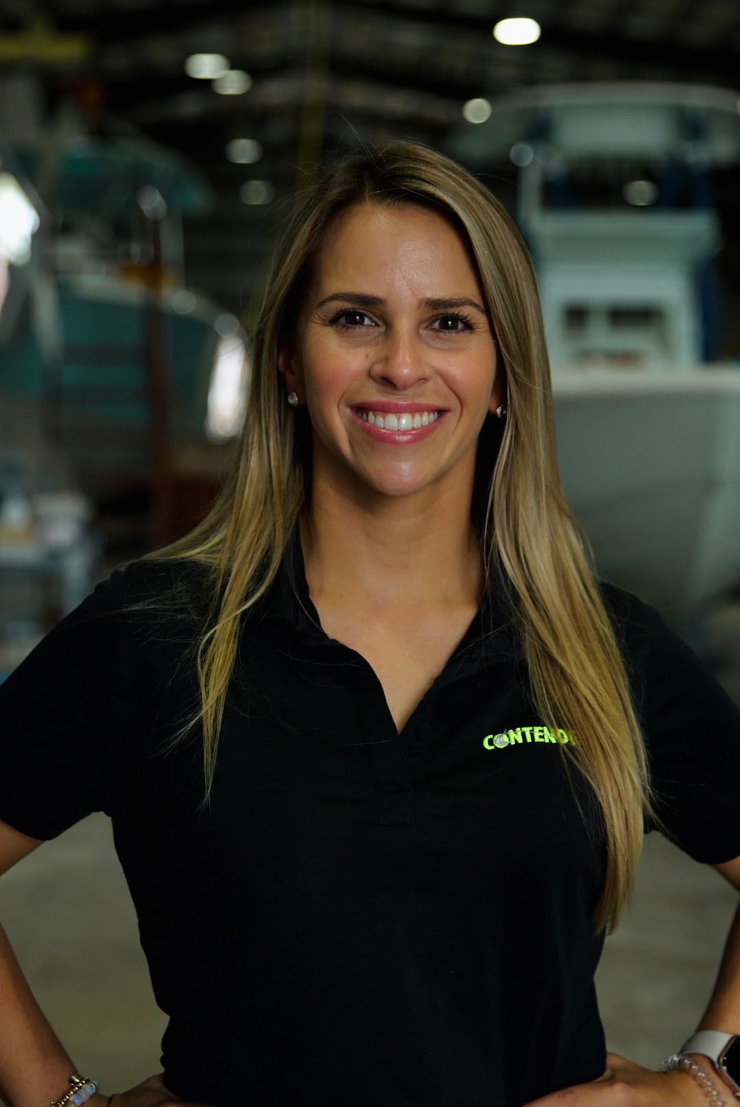 A woman with long blonde hair wearing a black polo shirt with a "Contender" logo stands indoors, smiling at the camera.