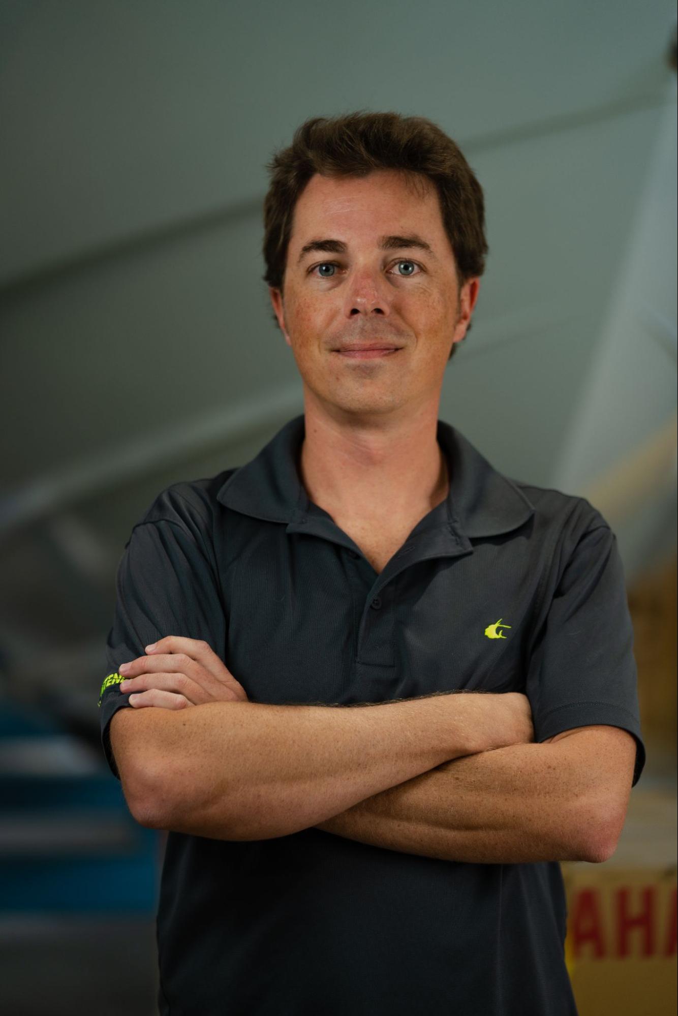 A person with short brown hair, wearing a dark grey polo shirt, stands with arms crossed against a blurred background.
