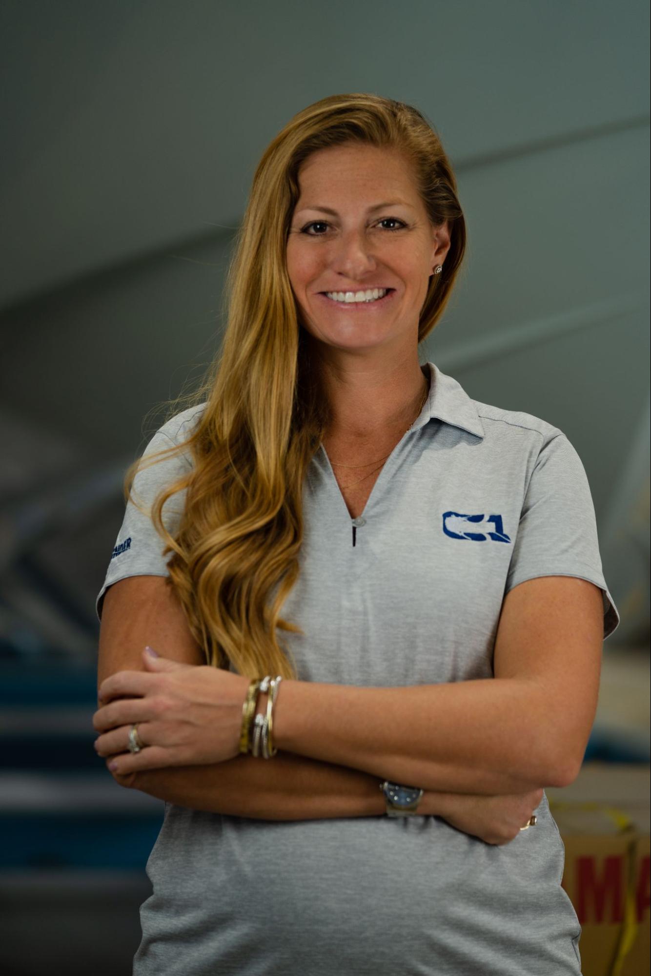 A woman with long hair smiles, arms crossed, wearing a gray polo shirt with logos. The background is blurry, possibly an indoor setting.
