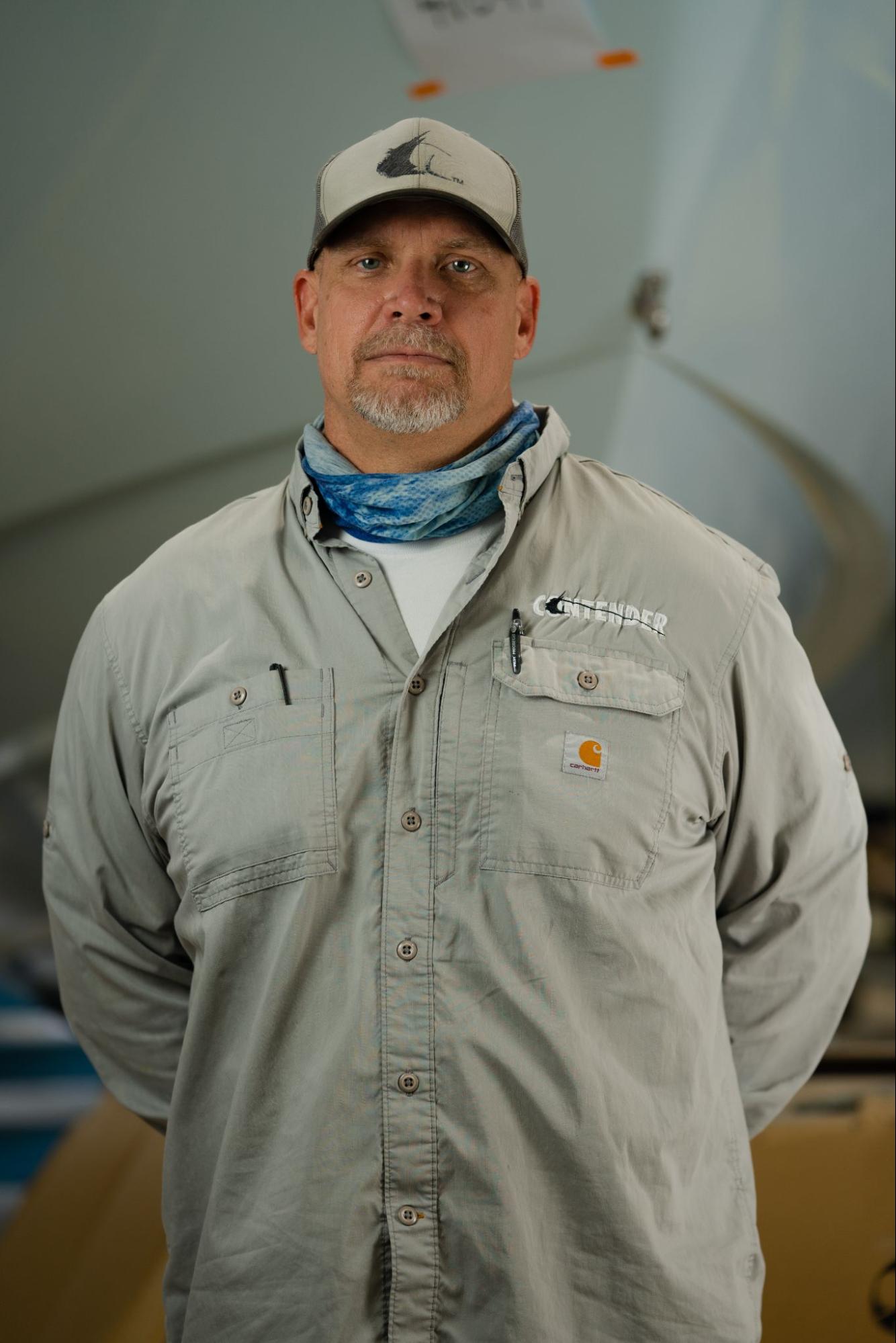 A man wearing a beige button-up shirt, a grey cap, and a blue neck gaiter stands in front of a blurred, industrial background.