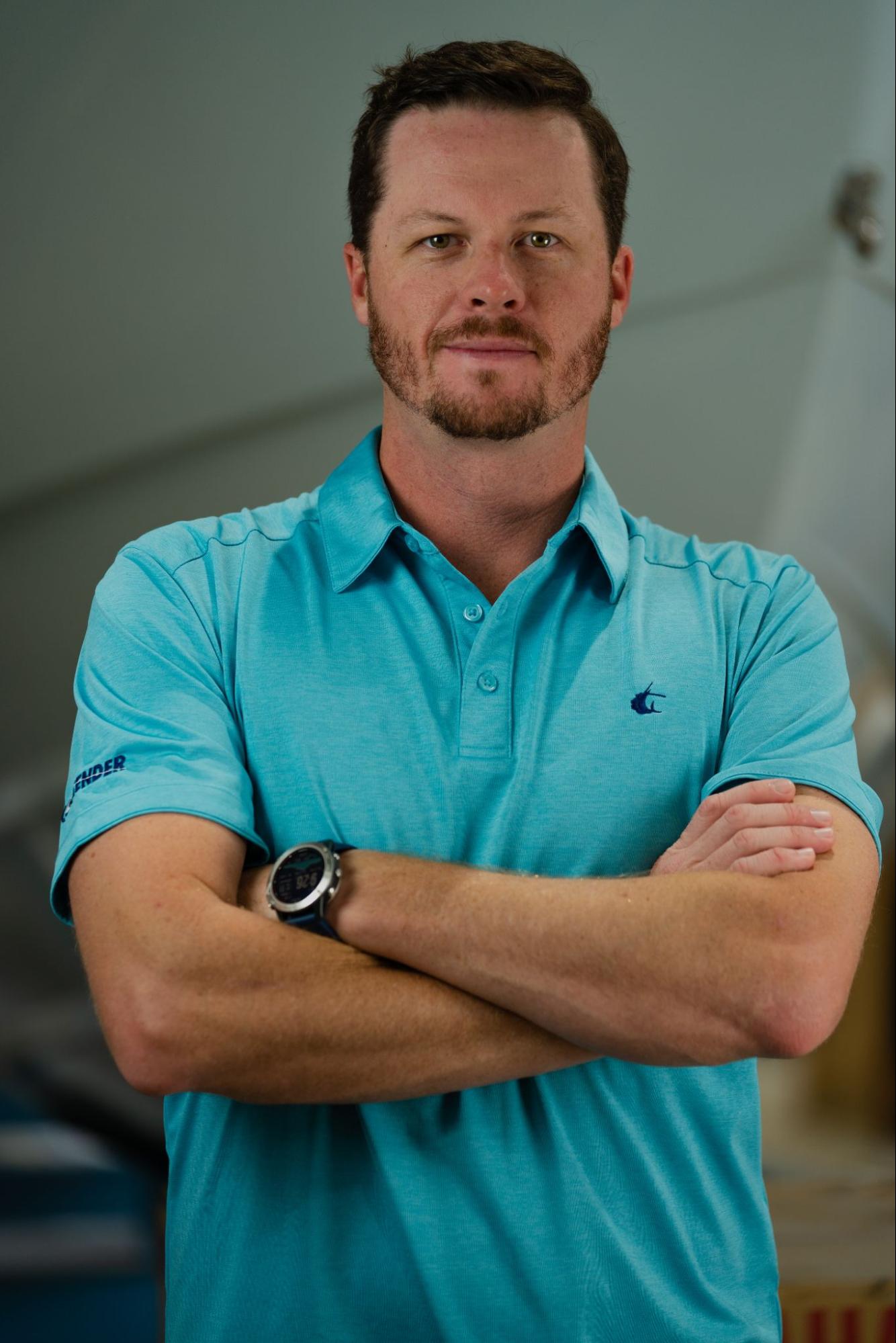 A man with a beard and short hair wearing a light blue polo shirt stands with his arms crossed.