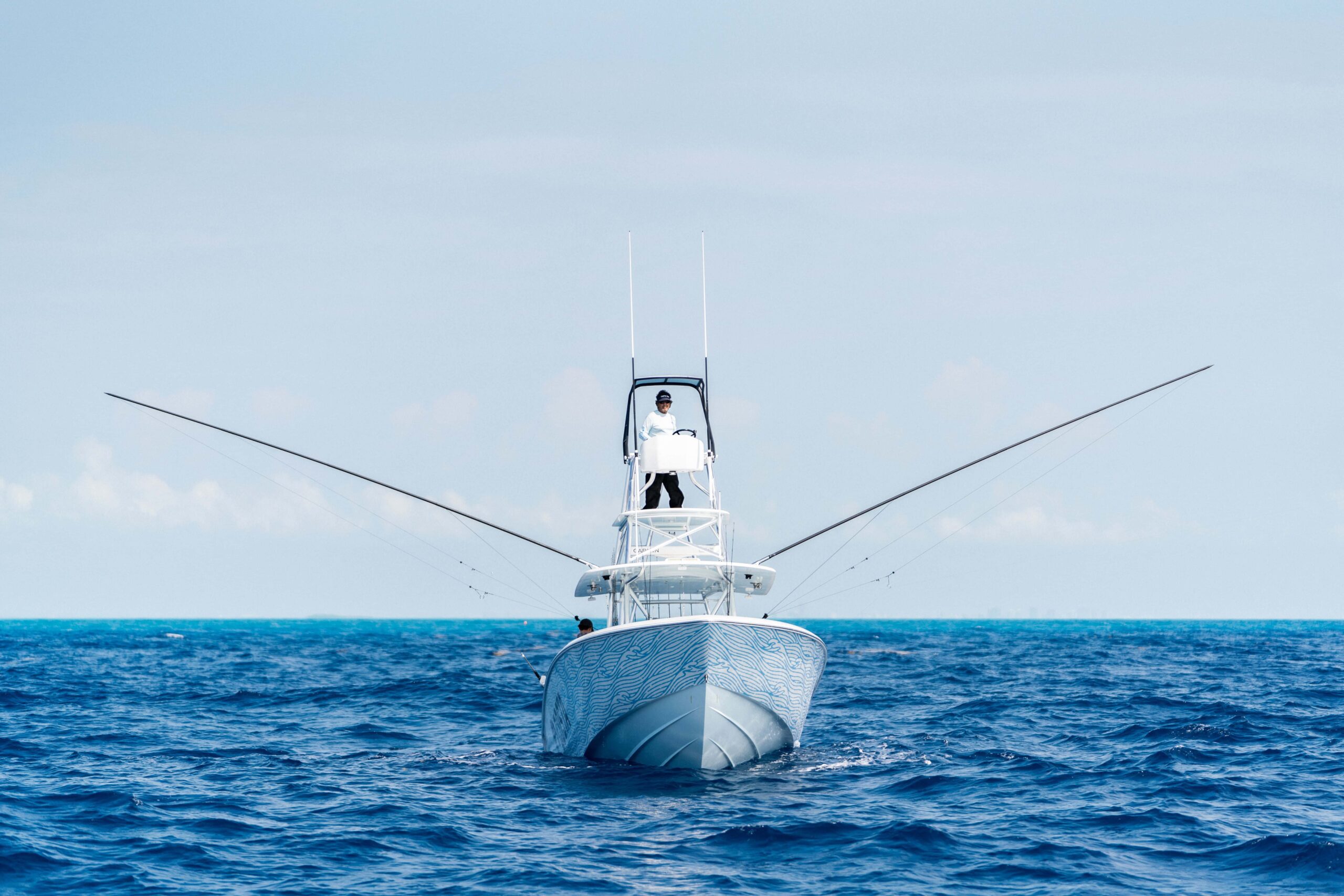 A fishing boat with extended outriggers is shown sailing on a calm ocean, with a person standing on the upper deck. The sky is clear and blue.